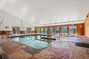 An indoor swimming pool with a hot tub, lounge chairs, and a view of the outdoors.