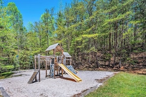 A wooden playground structure with a slide and swings, surrounded by a forested area.
