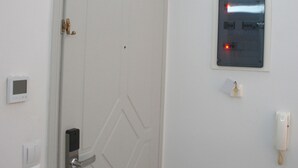 Een witte deur met een modern slot, een intercom bevestigd aan de muur en een plafondlamp.