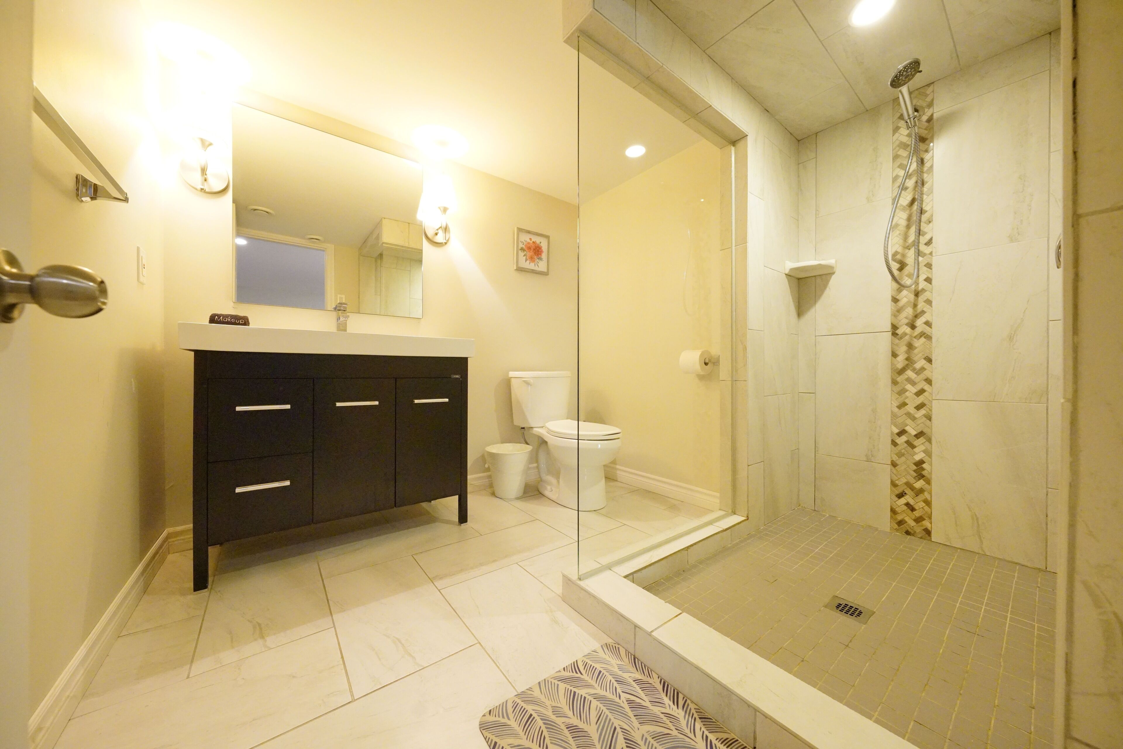 Un baño moderno con ducha envuelta en vidrio, tocador de color oscuro con espejo y retrete.