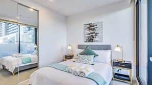 Quarto de hotel moderno com uma cama grande, duas mesas de cabeceira com luminárias, vista da cidade através de uma parede de vidro e um quadro na parede.
