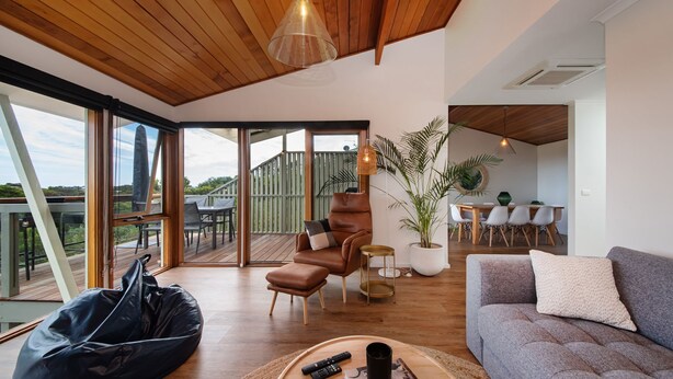 Un vaste espace de vie doté d’un plafond en bois, d’un fauteuil en cuir, d’un canapé gris et d’un coin repas à l’arrière-plan.