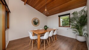 Un coin repas avec une table en bois et des chaises blanches, un luminaire suspendu, un miroir rond et une fenêtre donnant sur un paysage verdoyant.