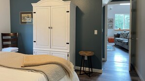 Una camera da letto con un letto, un armadio bianco, uno sgabello e una porta che conduce a un'altra stanza.