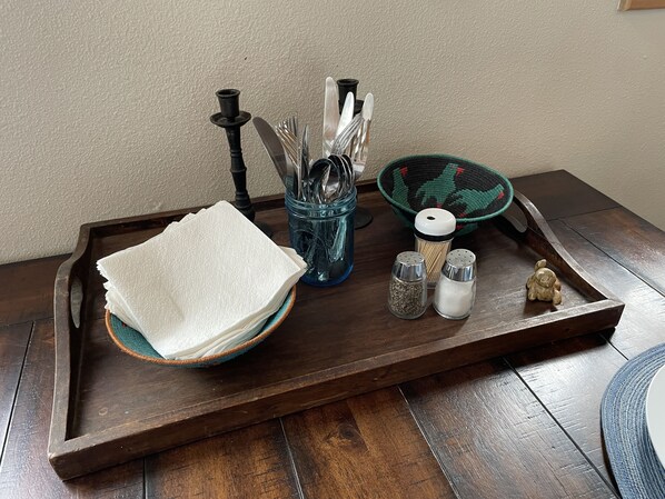 A wooden tray with a stack of white napkins, a blue glass jar with utensils, a green and black woven bowl, two salt shakers, and a small bear figurine.