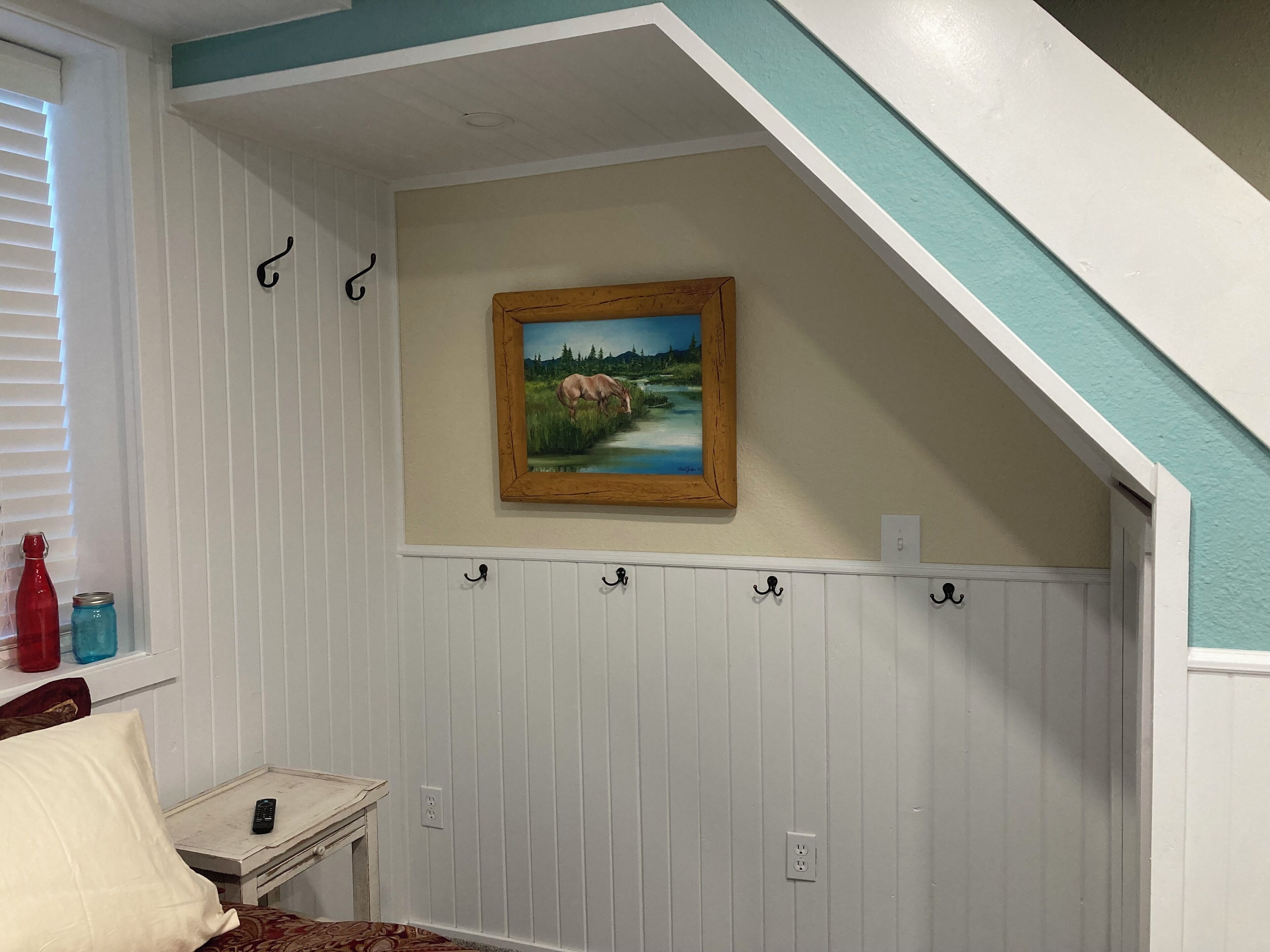 A room with a sloped ceiling, a painting of a landscape, a small table with a remote control, and a window with blinds.