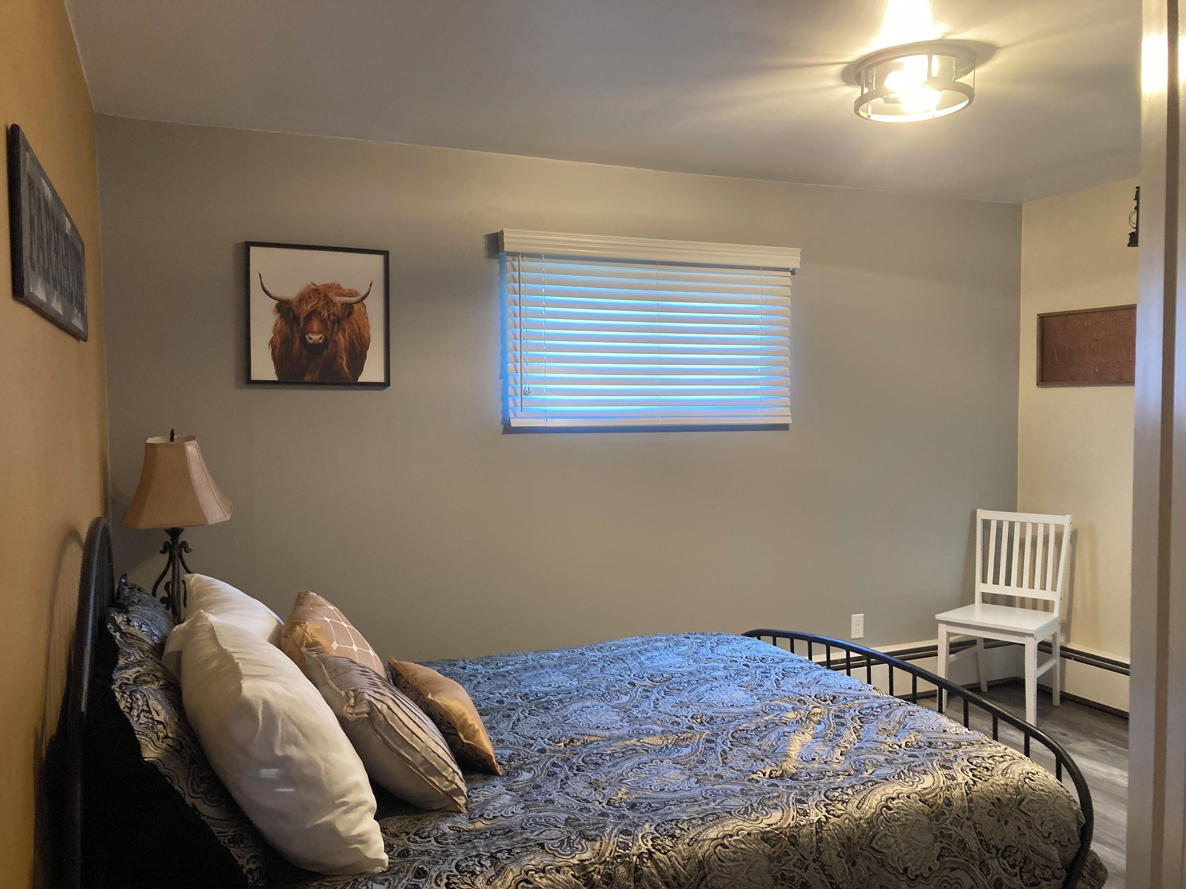 A bedroom with a bed, a chair, a lamp, a window with blinds, and a framed picture on the wall.