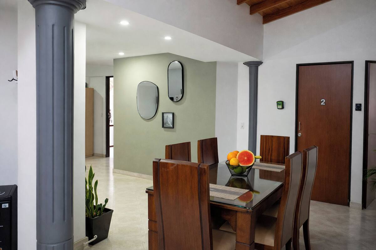 A dining area with a glass table, wooden chairs, a fruit bowl, and a potted plant.
