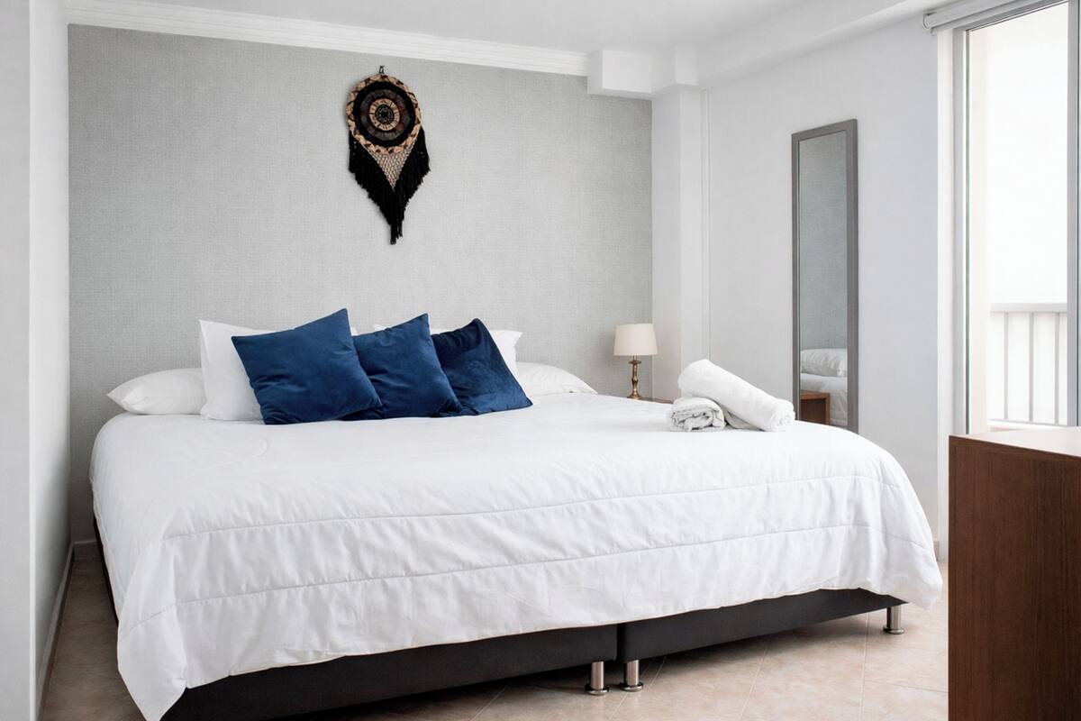 A hotel room with a large bed, a bedside table with a lamp, a mirror, and a wall-mounted decorative piece.