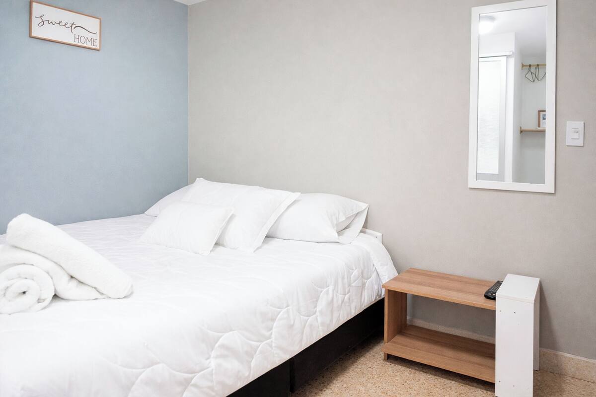 A neatly made bed with white linens and pillows, a small wooden nightstand with a remote control, a wall-mounted mirror, and a 'sweet home' sign on the wall.