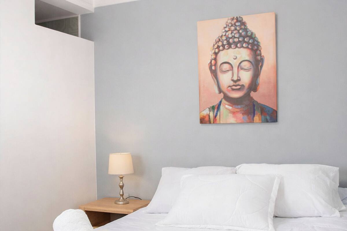 A bedroom with a bed, white pillows, a nightstand with a lamp, and a wall painting.