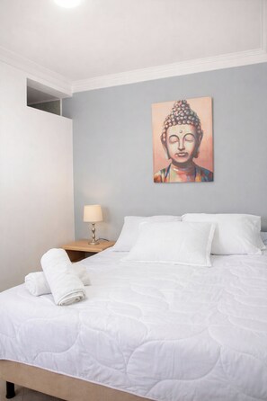 A neatly made bed with white linens, a wall-mounted painting, and a bedside table with a lamp. - Compostela Estadio (Medellín)