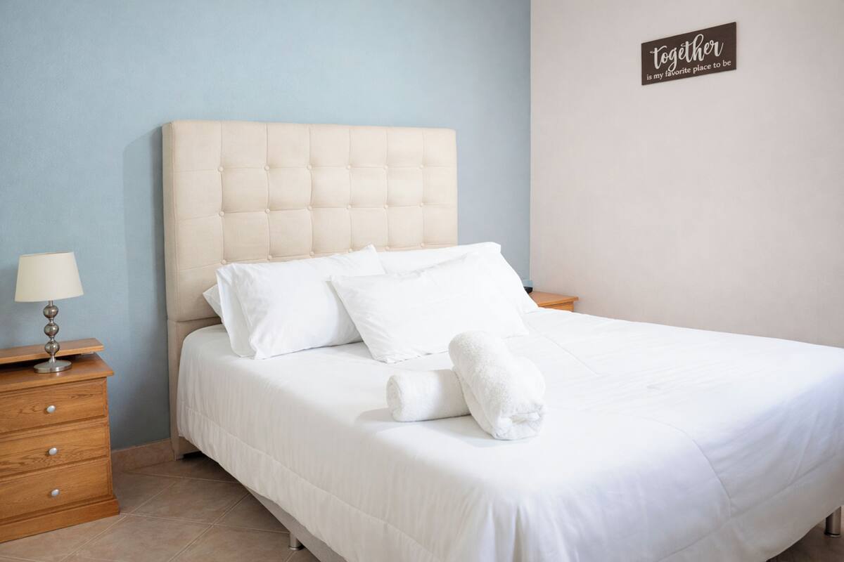 A neatly made bed with white linens and pillows, a wooden nightstand with a lamp, and a sign on the wall that reads 'together is my favorite place to be.'