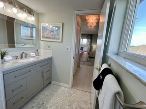 A modern bathroom with a large mirror, vanity, and a view of a bedroom through an open door. - Oceanfront Penthouse Suite, 2 King Beds, Exquisite Beach Views, Newly Renovated (Fernandina Beach)