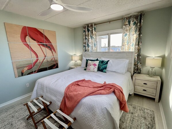 Une chambre avec un grand lit, deux tables de chevet, un ventilateur de plafond, une fenêtre avec des rideaux et une œuvre d’art murale.