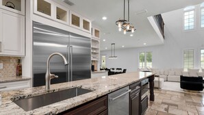 A modern kitchen with a stainless steel refrigerator, granite countertop, and a dining area with a sofa and table. - Mini Golf, Splash Pad, Indoor Playground, Movie Theatre & Summer Kitchen (Kissimmee)