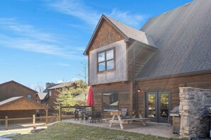 A two-story wooden building with a patio area featuring a grill, tables, and chairs.