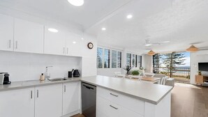 A modern kitchen with white cabinetry, a dishwasher, and a dining area with a view of the ocean.