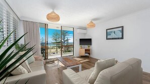 A modern living room with a sofa, a coffee table, and a TV. There is a large window with a view of trees and a building.