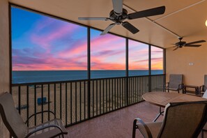 A balcony with a view of the ocean at sunset, a ceiling fan, and a table with chairs.
