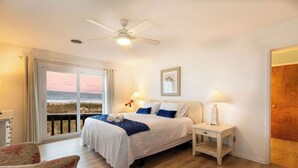 Ein Schlafzimmer mit Bett, Nachttischen, einem Sessel, einem Deckenventilator und einem großen Fenster mit Meerblick.