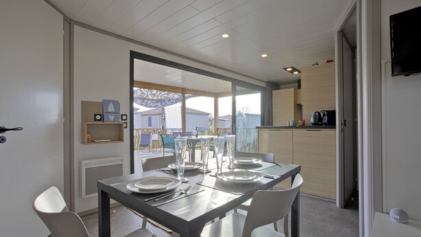 Un coin repas avec une table dressée pour quatre personnes, une cuisine visible à travers une porte ouverte et une vue sur un quartier résidentiel.