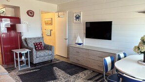 A compact living space with a sofa, a small table, a TV mounted on the wall, and a dining area with chairs. - Retro Ocean Front, Private Deck, Pool, Elevator, Linens  (Wildwood Crest)