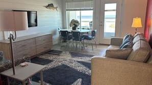 A living room with a sofa, a coffee table, a TV, a dining area with chairs, and a view of the ocean. - Retro Ocean Front, Private Deck, Pool, Elevator, Linens  (Wildwood Crest)