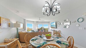 Un coin repas avec une table en verre, des chaises en bois et un lustre. De grandes fenêtres offrent une vue sur l’océan.