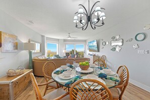 A dining area with a glass table, wooden chairs, and a chandelier. There is a view of the ocean through large windows.