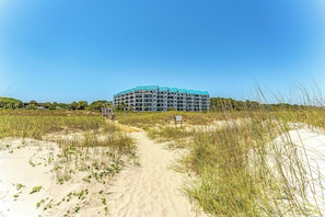 A beachfront hotel with a sandy path leading to the entrance, surrounded by tall grasses and a clear blue sky. - Remodeled Oceanfront Condo | Direct Balcony Views, Private Pool, King Suite (Hilton Head Island)