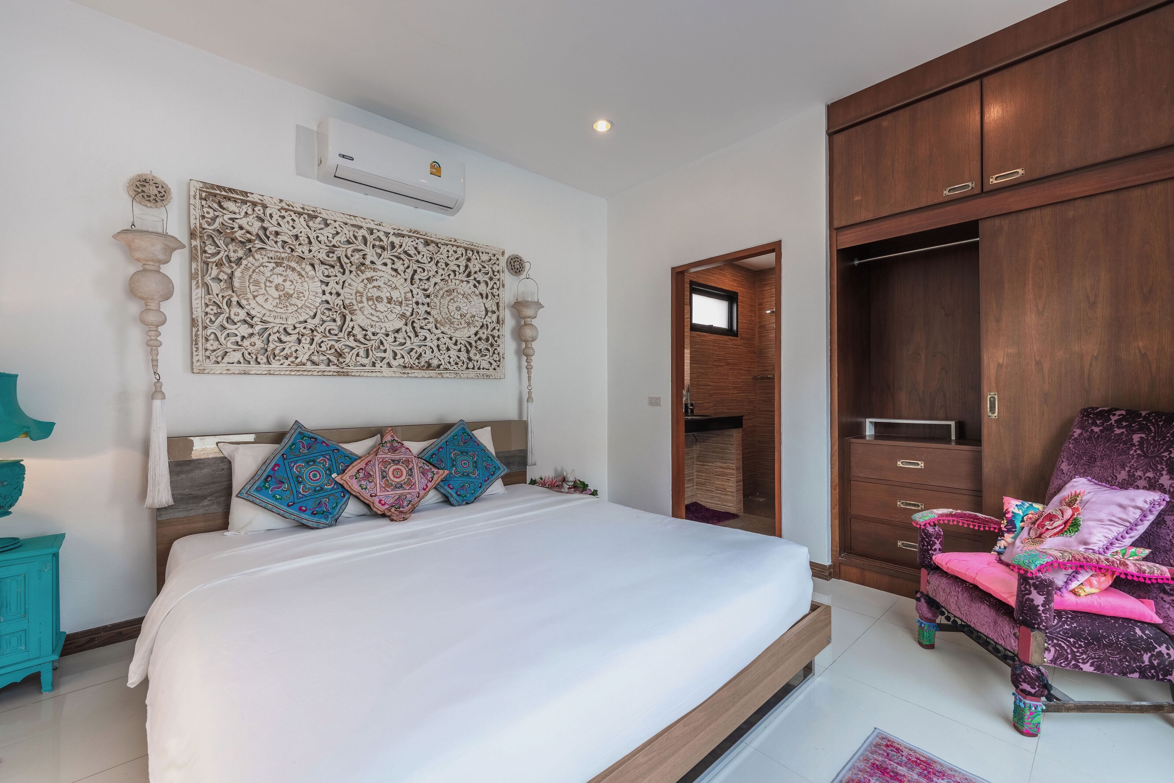 A bedroom with a large bed, a wooden wardrobe, and a chair with colorful cushions.
