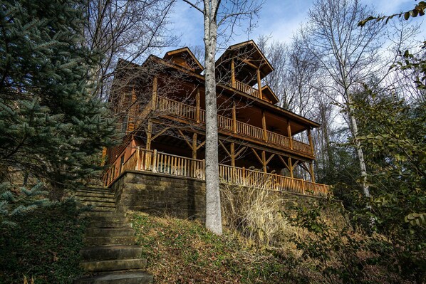 A multi-story wooden cabin with a stone staircase leading up to it, surrounded by trees and shrubs.