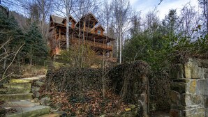 Una cabaña de madera rústica con varios balcones, rodeada de un bosque denso y escalones de piedra que llevan a ella.