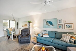 Een woonkamer met een blauwe bank, een lederen fauteuil, een eettafel met stoelen en een plafondventilator.