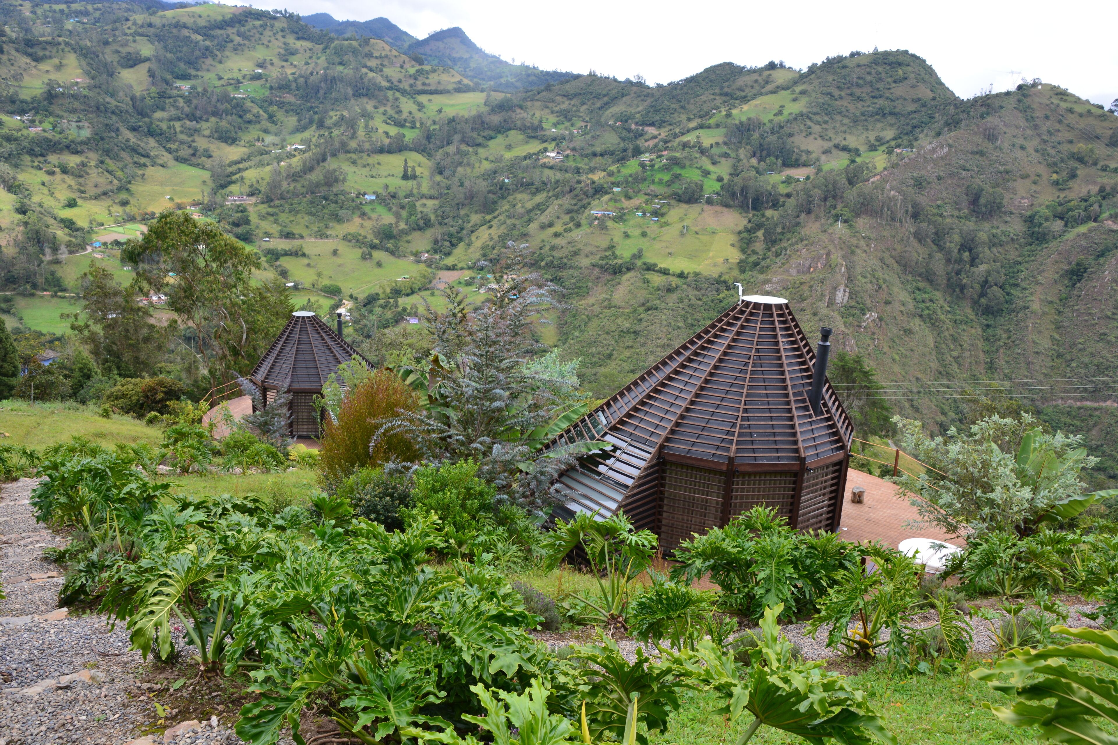 A traditional wooden house with a sloped roof, surrounded by lush greenery and a hilly landscape.