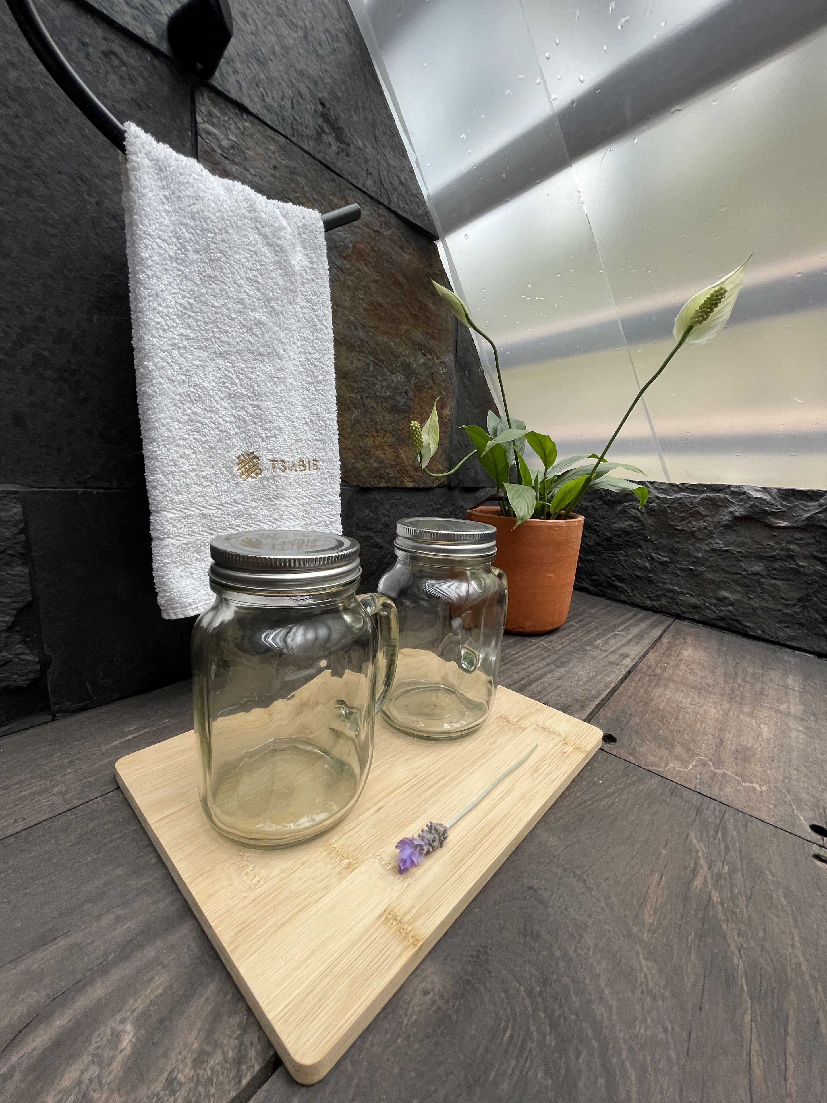 Dos frascos de vidrio sobre una tabla de madera, una toalla blanca con texto dorado y una planta en maceta.