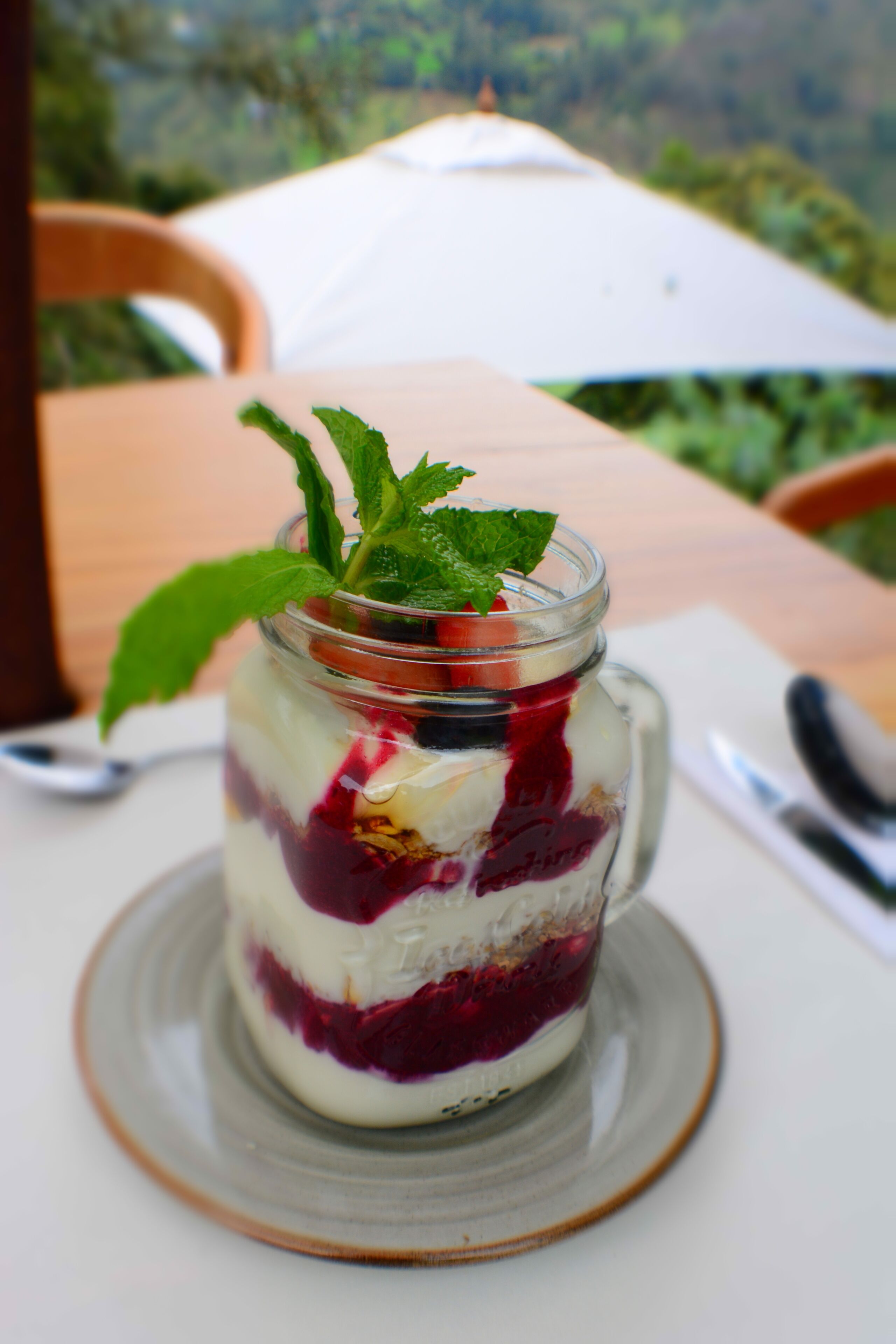 A layered dessert in a glass jar on a ceramic plate, with a spoon and napkin nearby.