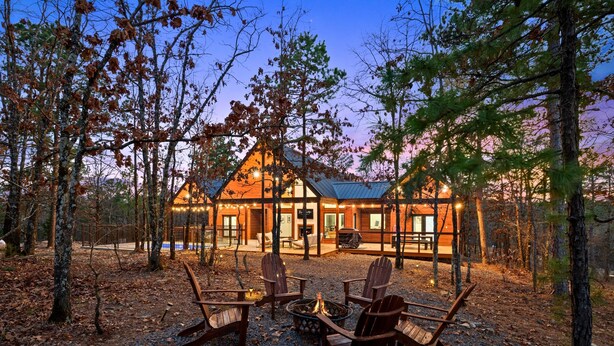 A lodge with a fire pit and Adirondack chairs in the foreground, surrounded by trees. - Luxe Pool Retreat for Couples and Small Groups (Broken Bow)