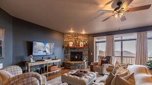 A cozy living room with a stone fireplace, a television mounted on the wall, and a ceiling fan. - Sugar Shack - Condo with amazing views, close to slopes! (Banner Elk)