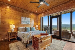 A wooden cabin bedroom with a bed, bedside lamps, a wooden chest, and a large sliding glass door leading to an outdoor patio with seating. - Fine As Wine ~ Resort Pool, Swim-Up Bar, Pickleball, Putt-Putt & Epic Views (Sevierville)