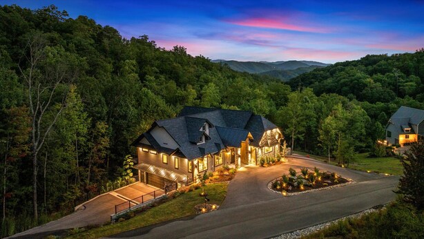 A large, multi-story house with a stone facade and a steeply pitched roof, surrounded by dense forest.
