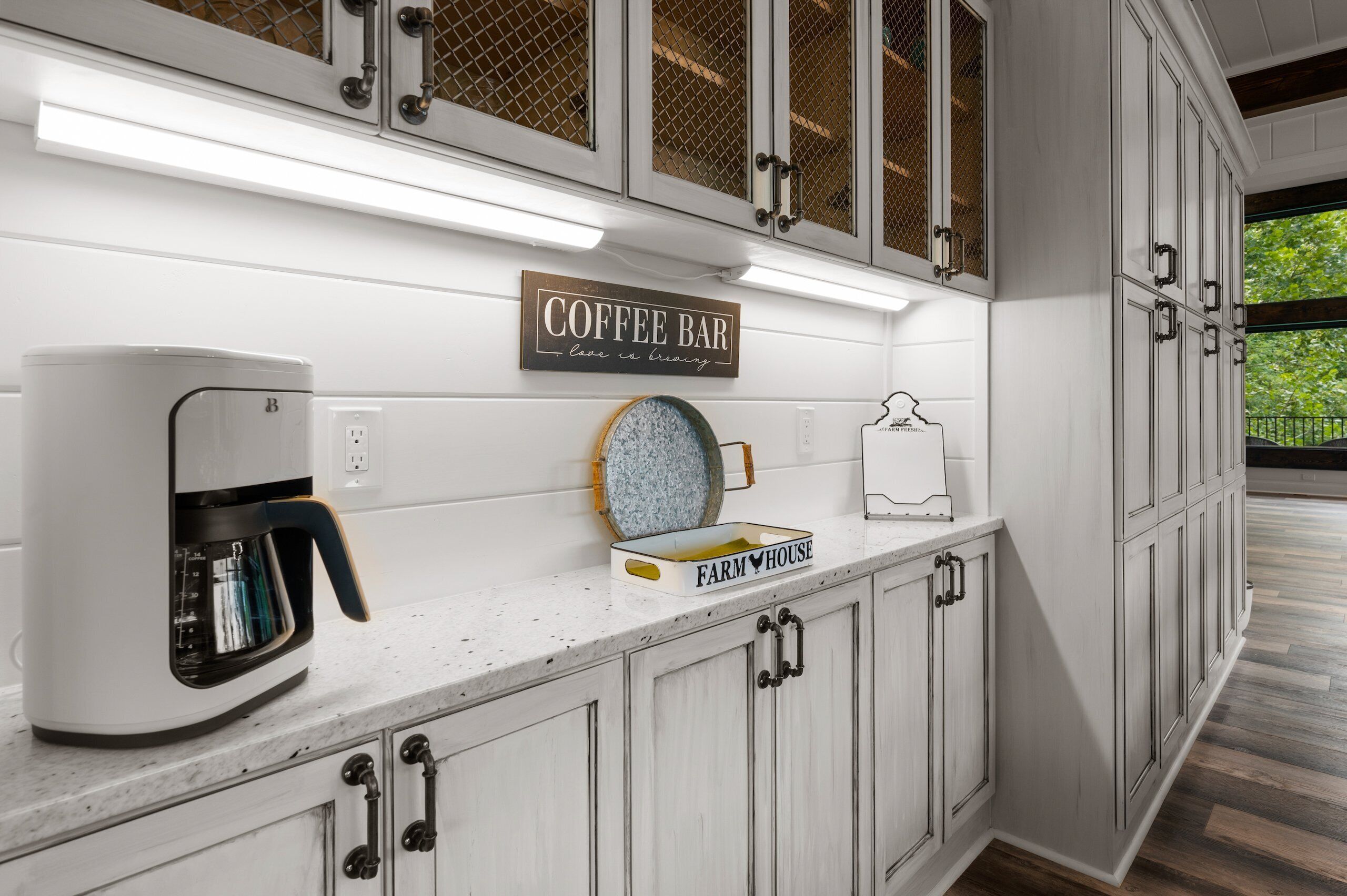 Une cuisine avec des meubles blancs, une machine à café, un panneau décoratif de style rustique et une fenêtre donnant sur les arbres.