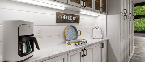 Une cuisine avec des meubles blancs, une machine à café, un panneau décoratif de style rustique et une fenêtre donnant sur les arbres.