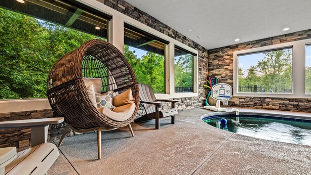 An indoor pool area with a stone wall, large windows, and a wicker chair. - Firefly Mtn Retreat ~ Luxury 4BR Pool Cabin in The Summit at Bluff Mountain (Sevierville)