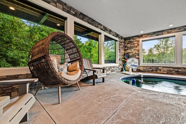 An indoor pool area with a stone wall, large windows, and a wicker chair. - Firefly Mtn Retreat ~ Luxury 4BR Pool Cabin in The Summit at Bluff Mountain (Sevierville)