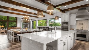 A spacious kitchen with a large island, white cabinetry, and a stone backsplash. There are multiple hanging lights and a large window providing natural light. - Firefly Mtn Retreat ~ Luxury 4BR Pool Cabin in The Summit at Bluff Mountain (Sevierville)