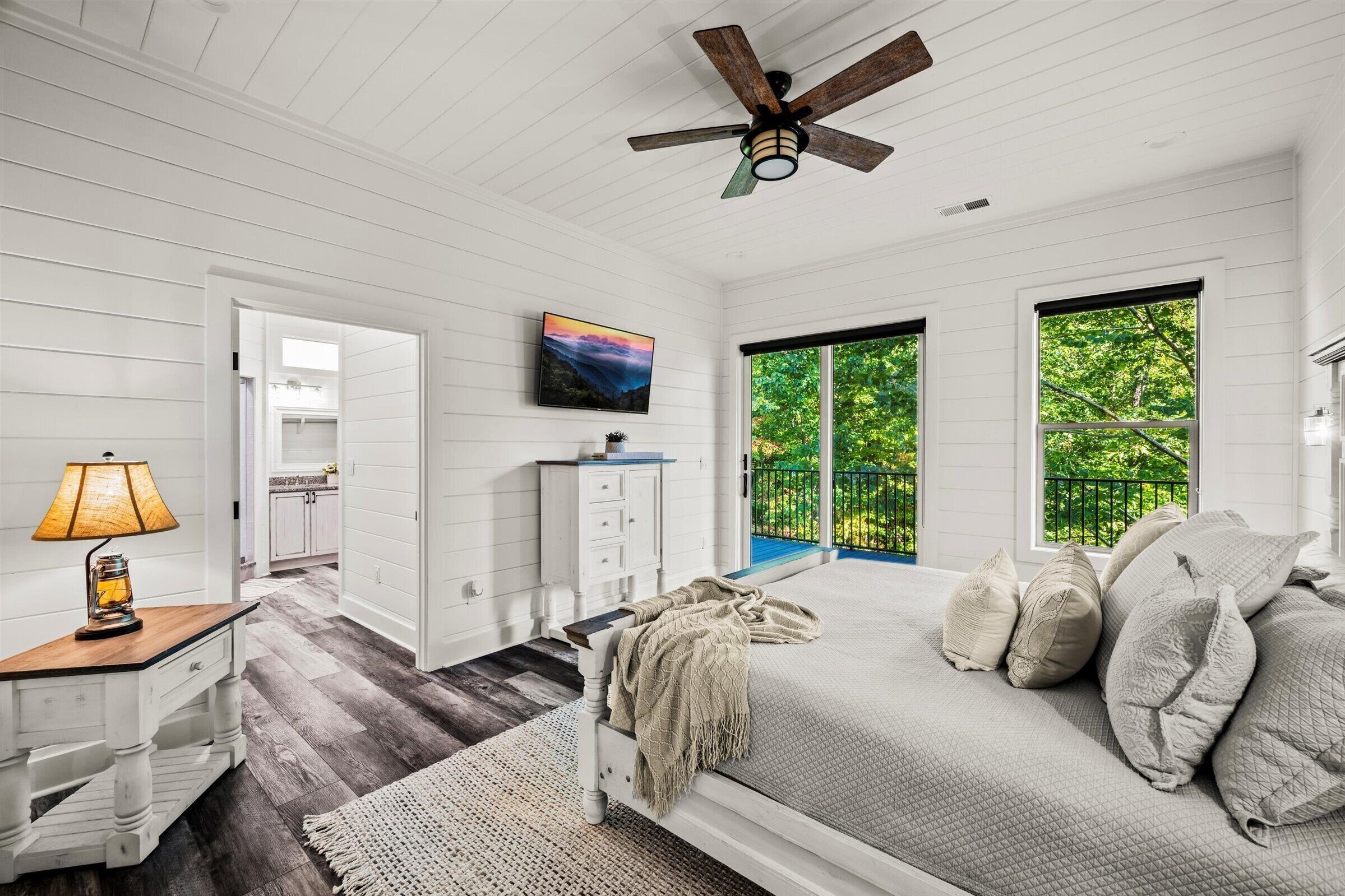 A bedroom with a bed, a nightstand, a TV, a ceiling fan, and a view of the outdoors.