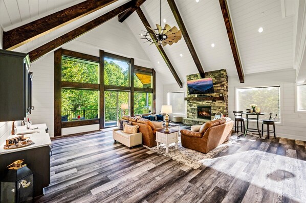 A spacious living room with a high ceiling, large windows, a fireplace, and a dining area. - Firefly Mtn Retreat ~ Luxury 4BR Pool Cabin in The Summit at Bluff Mountain (Sevierville)