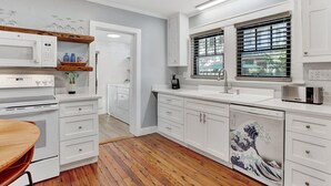 Una cucina moderna con mobili bianchi, pavimento in legno e vista sull'esterno attraverso una finestra con persiane.